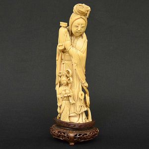 <b>Estatueta</b> chinesa em marfim representando figura de anciã com jovem, ambas com atributos às mãos. Base em madeira entalhada. Séc. XIX. Falta um pé da base de madeira. Alt. total 26 cm