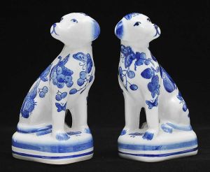 <B>Par de cães </b>sentados, em porcelana, decorados com frutos em azul sobre fundo branco. Alt. 21 cm