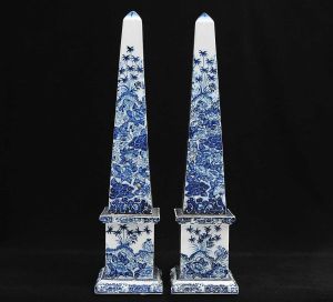<b>Par de obeliscos</b> em porcelana, decorados em azul com palmeiras, flores e figuras de anjos sobre fundo branco. Alt. 63,5 cm